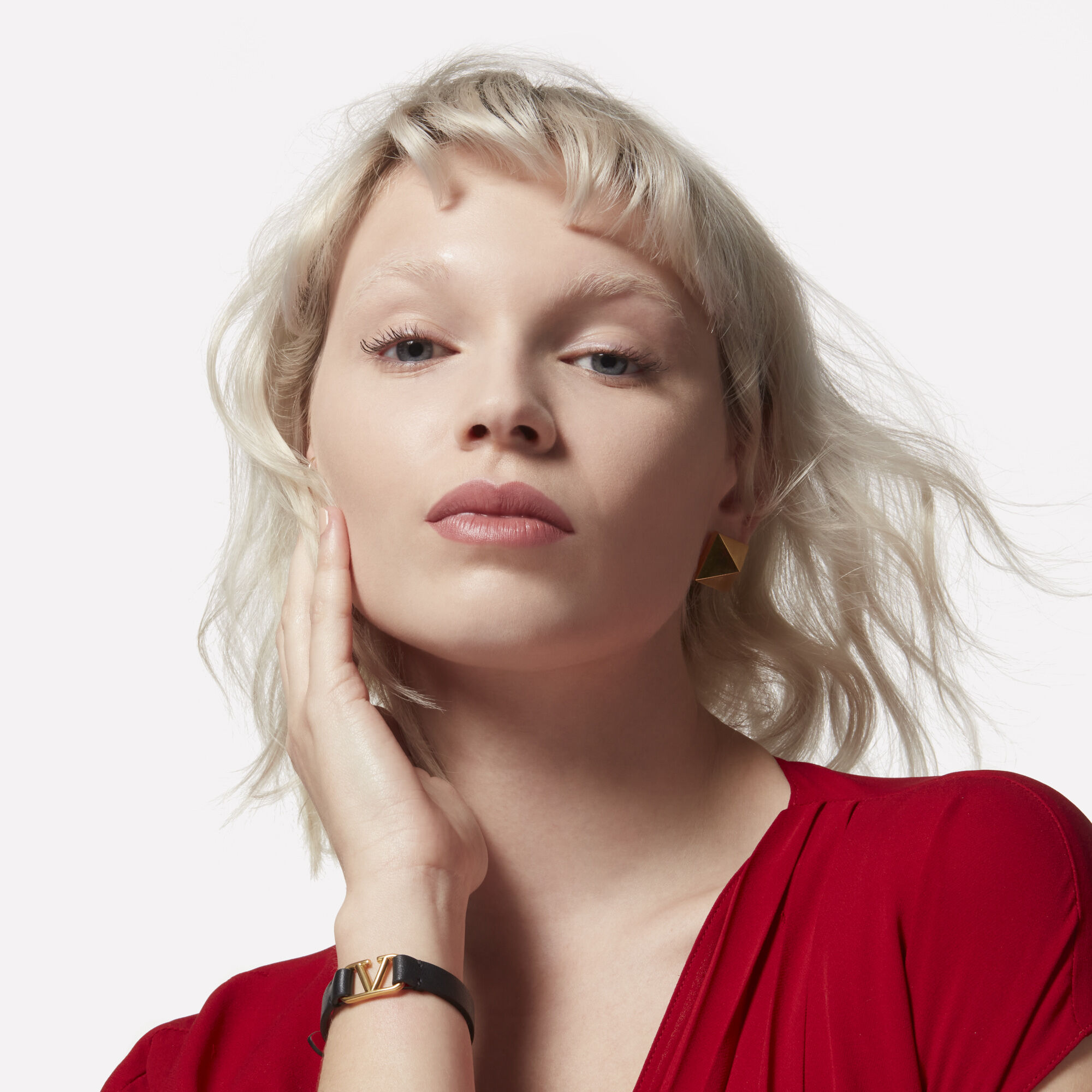 A model with platinum blonde hair caressing her face wearing a Valentino Beauty logo bracelet